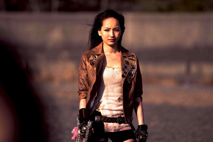 In the movie scene SchoolGirl Apocalypse 2011, Higarino with a fierce look standing, hands down, holding her sword with her right hand, has black long hair and scars on left cheek wearing black shorts, black gloves, black belt, pink doll keychain and brown leather jacket over a white tank top.