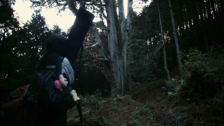 In the movie scene SchoolGirl Apocalypse 2011, Higarino stands in the middle of a forest looking at the tree in middle, has long black hair with a black shoulder bag with one pink and two yellow doll keychains while holding her sword covered in black cloth, wearing a white seifuku school uniform with a black marine collar, skirt, and socks.