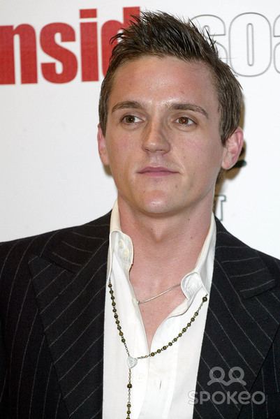 Scott Neal with a tight-lipped smile and looking afar during the Inside Soap Awards in London England while wearing necklaces and a white long sleeve under a black striped coat