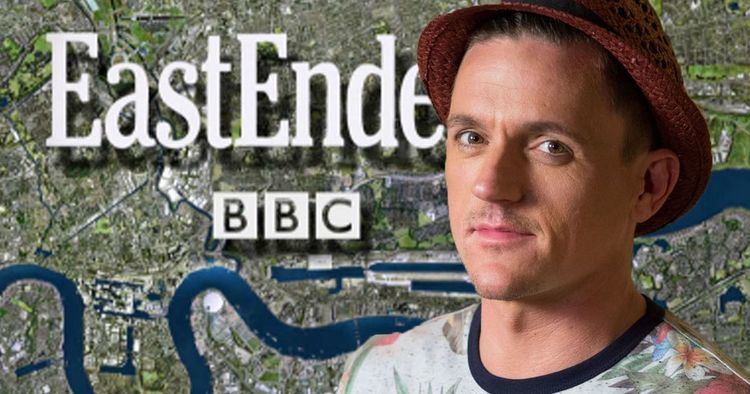 Scott Neal smiling with a map in the background and a caption "EastEnders" and "BBC". Scott is wearing a floral shirt and a brown knitted hat