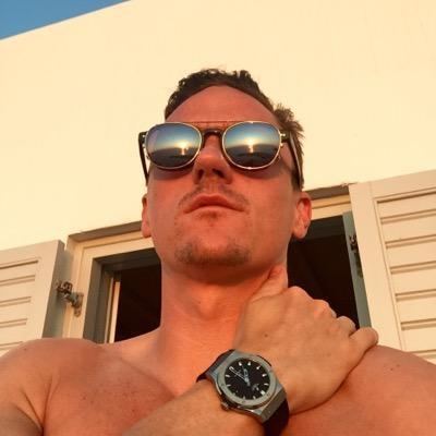 Scott Neal looking afar with a serious face, mustache, beard, hand on his shoulder, and topless while wearing sunglasses and a wristwatch