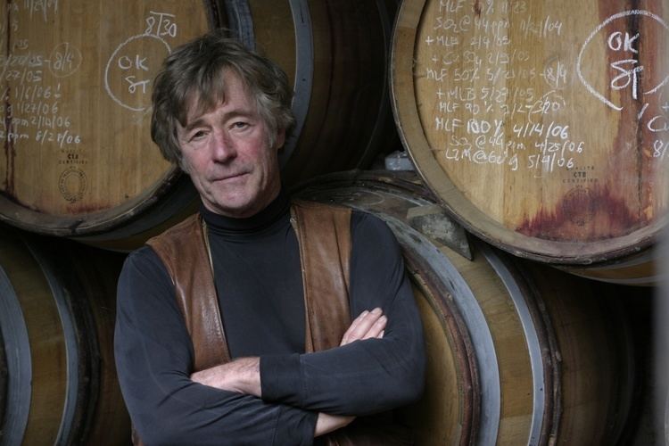 Sean Thackrey is serious, has light brown hair, wearing a brown and black jacket both hands crossed, standing inside his small Bolinas winery.
