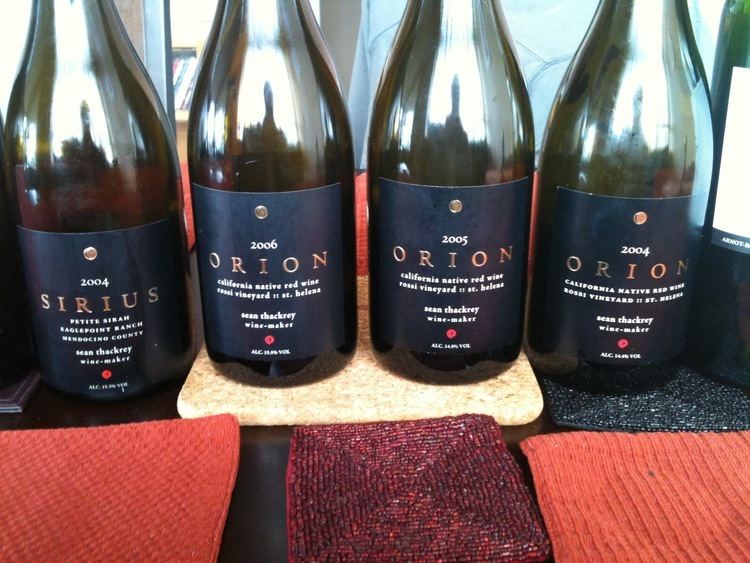 On left is a Sirius wine (2004) has a black sticker with the information. 2nd from left is an Orion wine (2006) that has a black sticker with the information. 3rd from left is an Orion wine (2005) that has a black sticker with the information. On (right) is an Orion wine (2004) that has a black sticker with the information on the table with different colors of table cloth.