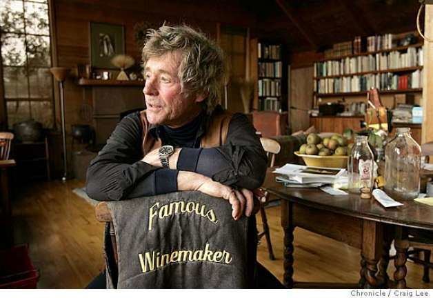 Sean Thackrey is serious, has white hair, sitting down on a brown chair with a cloth with a print/design “Famous Winemaker” inside his art gallery, left hand wearing a silver wristwatch wearing a brown and black jacket. He has a table with papers and bottles and fruits.