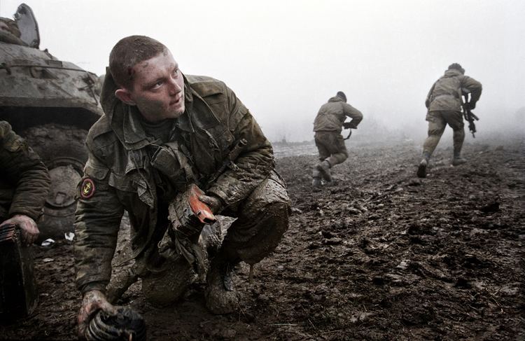 A soldier kneeling on the ground while looking afar and the two soldiers at his back are running away while holding their guns during the Second Chechen War