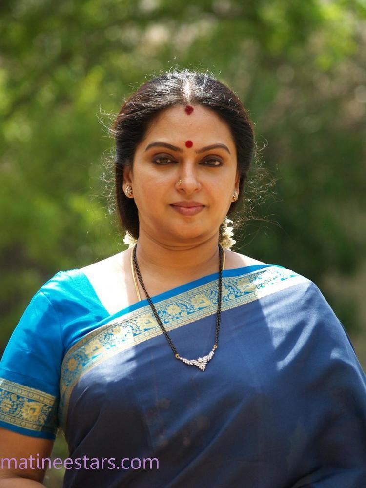 Seetha with a serious face, wearing a necklace, earrings, and a blue dress.
