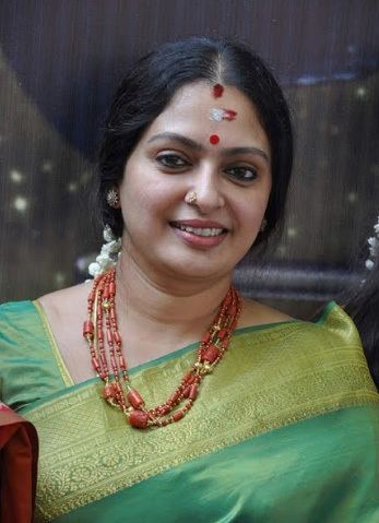 Seetha smiling, with black hair, wearing earrings, red necklace, and a green dress.