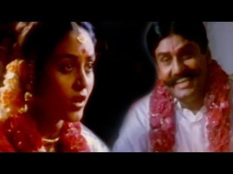 In the movie scene of Seevalaperi Pandi 1994, Saranya Ponvannan (left) is sad, mouth half open remembering the image of Napoleon (right), she has black hair and bindi on her forehead wearing a gold accessory over her head a pair of earrings, piercing on her nose and a flowers around around her neck, on the right Napoleon is smiling mouth half open, has mustache and black hair, wearing a white top and flowers around his neck.