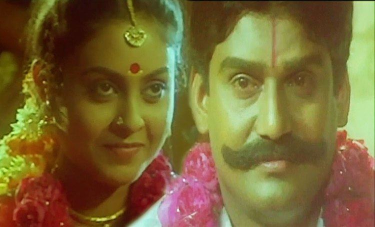 In the movie scene of Seevalaperi Pandi 1994, Saranya Ponvannan (left) is smiling remembering the image of Napoleon (right), she has black hair and bindi on her forehead wearing a gold accessory over her head a pair of earrings, piercing on her nose and a flowers around her neck, on the right Napoleon is smiling, has red line on his forehead, mustache and black hair, wearing a white top and flowers around his neck.