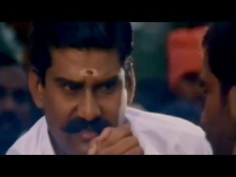 In the movie scene of Seevalaperi Pandi 1994, from right Napoleon is serious looking at his opponent while doing an arm wrestling using his right arm, has bindi on his forehead, a mustache and black hair, wearing white long sleeve, on the right a man facing Napoleon in arm wrestling using his right arm, has black hair.