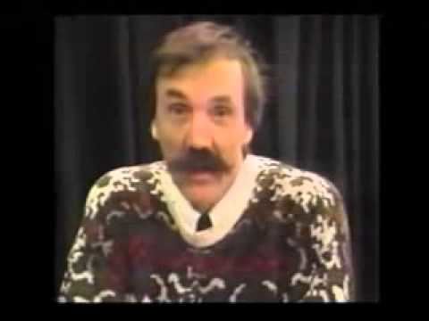 Serge Monast is serious, has black hair, and a mustache, wearing a white sweater with a brown and red design.