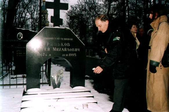 Donald Jackson putting a flower on the gravesite of Sergei Grinkov and behind him is a woman wearing a brown trench coat while Donald is wearing a black jacket and pants