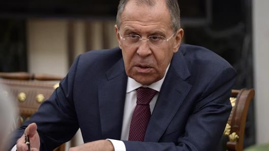 Sergey Lavrov with a serious face while looking at something and holding a black pen, wearing eyeglasses, a blue coat over white long sleeves, and a maroon necktie.