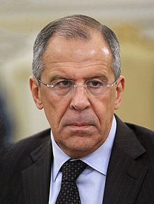 Sergey Lavrov with a serious face, wearing eyeglasses, a black coat over white long sleeves, and a black necktie.