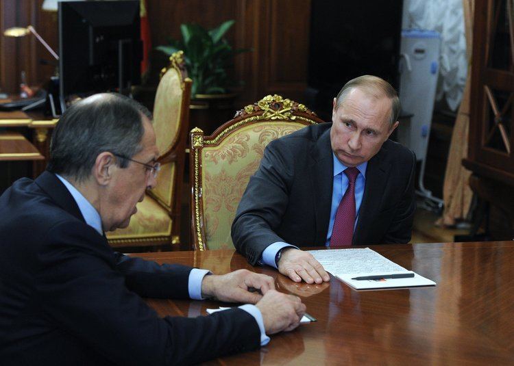Sergey Lavrov is talking while Vladimir Putin is looking at him. Sergey is wearing eyeglasses and a black coat over white long sleeves while Vladimir is wearing a black coat over blue long sleeves, and a red necktie.