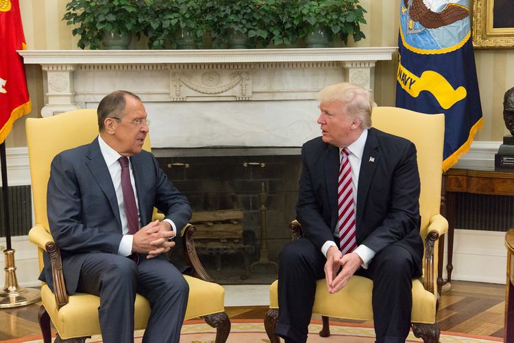 Sergey Lavrov and Donald Trump are talking while sitting on yellow chairs. Sergey is wearing eyeglasses, a gray coat over white long sleeves, a maroon necktie, gray pants while Donald is with blonde hair wearing a black coat over white long sleeves, a red striped tie, and black pants.