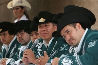 The man on the left is serious and wears silver earrings, a black hat with a gold design in the middle, and a white-green suit with a design on his chest. 2nd from left a man is serious, has a beard, and a mustache, and wears a black hat, and a white-green suit with a design on his chest, and on his right hand. 3rd from left a man is serious, wears a black hat, and a white-green suit. 4th from left is Sergio Gomez, smiling, has black hair, both hands holding the microphone, wears a black hat with a gold design in the middle, and a white-green suit with a design on his chest. 5th from left, a man is wearing a black hat and a white-green suit with a design on his chest. The man on the right is serious, has black hair, a beard, and a mustache, left hand on the table, and wears a black hat, and a white-green suit with a design on his left arm and hands. A woman (left) behind is serious, has black hair, wears silver earrings, a white hat with a design on left, and a white shirt.