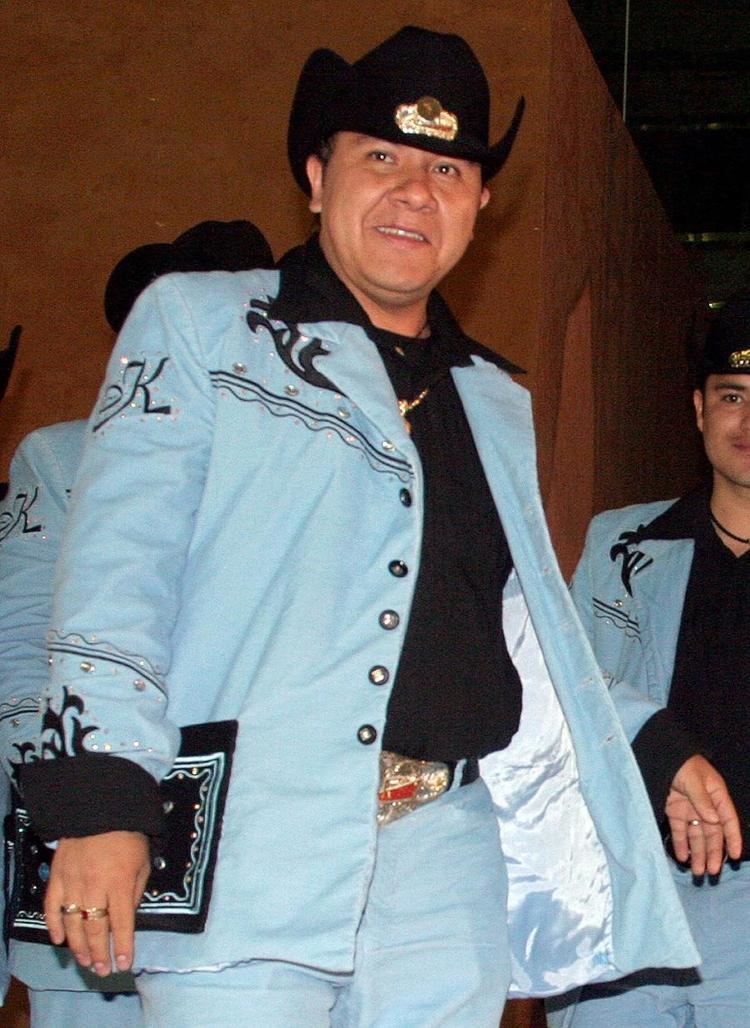 Sergio Gomez is smiling and wears a black hat with a gold design in the middle, a gold necklace, 2 gold rings on his right hand, and 1 gold ring on his left hand, a black shirt under a blue suit with a black design and silver beads on his right chest, a “K” print on his right arm with beads and on his hands, a black pocket with silver beads and blue design on right, a black belt with red-gold design in the middle and, blue pants. A man behind Sergio is wearing a black hat and a blue suit with the print “K” on his right arm with beads. A man on the right is smiling, wearing a black necklace, a black hat with a gold design in the middle, a black shirt under a blue suit with a black design and silver beads on his right chest, and blue pants.