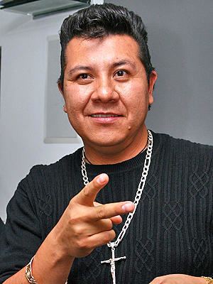 Sergio Gomez is smiling, has black hair and acne on his lower left cheek, and his right hand doing the peace sign, wears a silver bracelet on his right hand, a gold rosary necklace, and a black shirt.