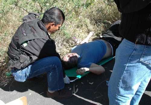 A man on the left has black hair, his right-hand holds the green spine board, and wears a black jacket under a black bulletproof suit with pockets, denim pants, and brown shoes. In the middle is Sergio Gomez, dead, has black hair, mouth half open, eyes closed, right hand up, lying on the green spine board, wearing a blue shirt, white underwear, and black pants. A man on the right is wearing a black jacket and blue pants.