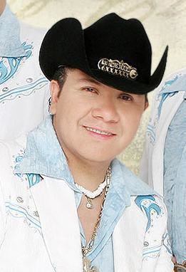 Sergio Gomez is smiling, has black hair, wears a white and gold necklace, a black hat with a gold design in the middle, and a denim-white jacket with a blue and silver beads design. Behind him are two people, wearing a denim-white jacket with blue and silver beads design.