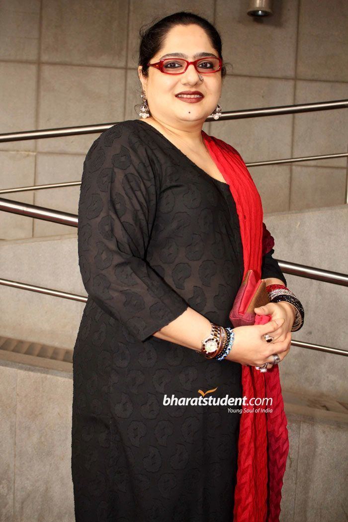 Shagufta Ali smiling, wearing eyeglasses, earrings, and a black dress with a red scarf.