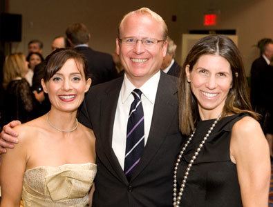 Catherine Hooper, Eric Swanson, and Shana Madoff smiling. Catherine is wearing a gold tube dress and a necklace, Eric holding Catherine's shoulder and wearing a white shirt under a black coat, a necktie with stripes, and eyeglasses while Shana is wearing a black sleeveless dress and a pearl necklace