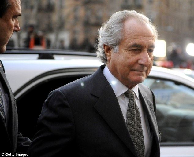 Bernie Madoff smiling while being accompanied by an official and wearing a white shirt under a black coat and a gray necktie