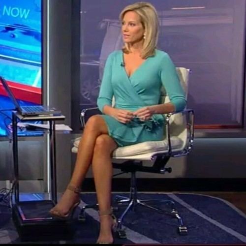 Shannon Bream sitting on a white office chair and looking at something, with a serious face and blonde shoulder-length hair, while wearing brown heels, earrings, and a wireless mic on her aqua-green long sleeve dress.
