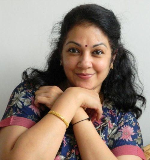 Shanthi Krishna smiling while both arms are crossed to each other and leaning his chin on her right hand, she has black hair, a nose piercing and a bindi on her forehead wearing earrings, bracelets on both wrists, and a floral blouse