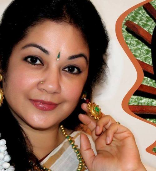 Shanthi Krishna smiling while touching her left earring with her left hand, she has black hair, a nose piercing and a bindi on her forehead wearing dangled earrings, a necklace, and a saree