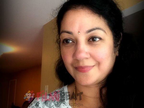 Shanthi Krishna smiling with her black hair, a nose piercing, and a bindi on her forehead wearing earrings, a gold necklace and printed shirt