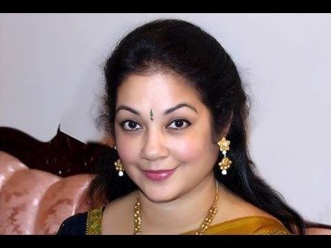 Shanthi Krishna smiling while sitting on a pink couch, she has black hair, a nose piercing and a bindi on her forehead wearing a dangled earrings, gold necklace, and a saree