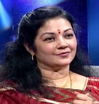 Shanthi Krishna smiling with her black hair, a nose piercing, and a bindi on her forehead wearing dangled earrings, a gold necklace, and a saree