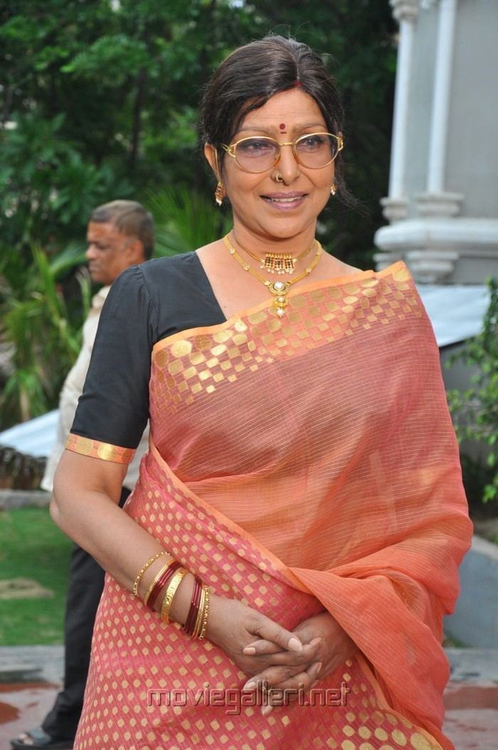Sharada wearing eyeglasses, gold jewelries and black and orange dress while her hands together