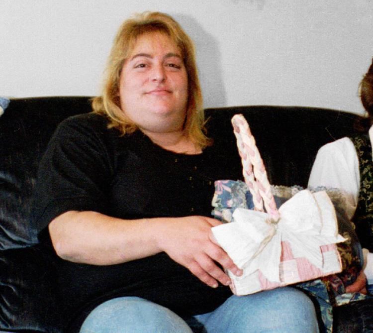 Sharon Lopatka with blonde hair, sitting on a black couch and wearing a black shirt while holding a basket.