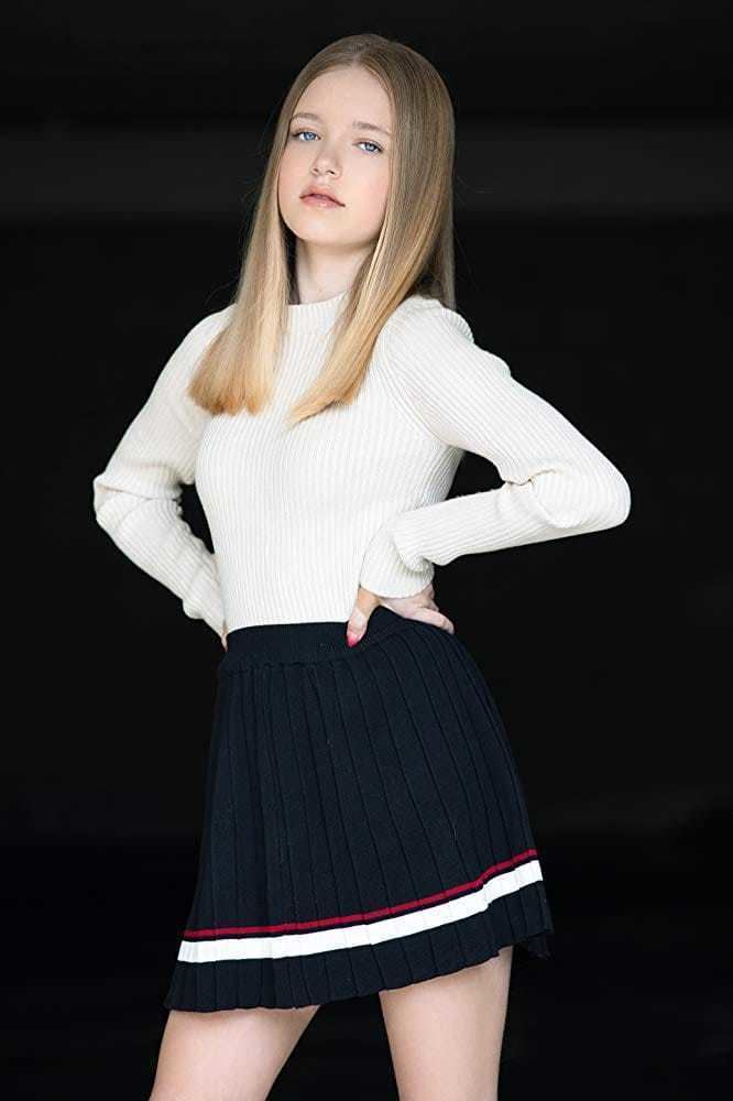 Shay Rudolph posing while hands on her waist and she is wearing a white long sleeve blouse and black skirt with white and red lines