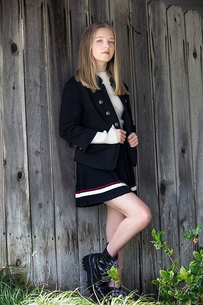 Shay Rudolph leaning on a wood fence while wearing a black coat, white long sleeve blouse, black skirt with white and red lines, and black shoes