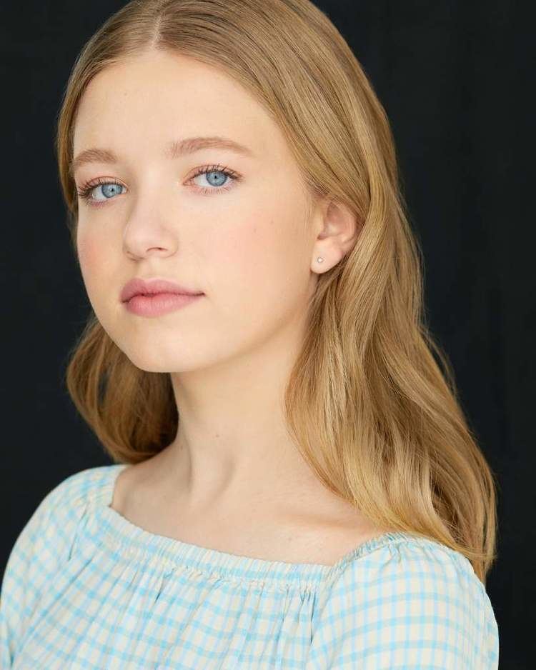 Shay Rudolph with blue eyes and blonde hair while wearing a light yellow and blue blouse and earrings