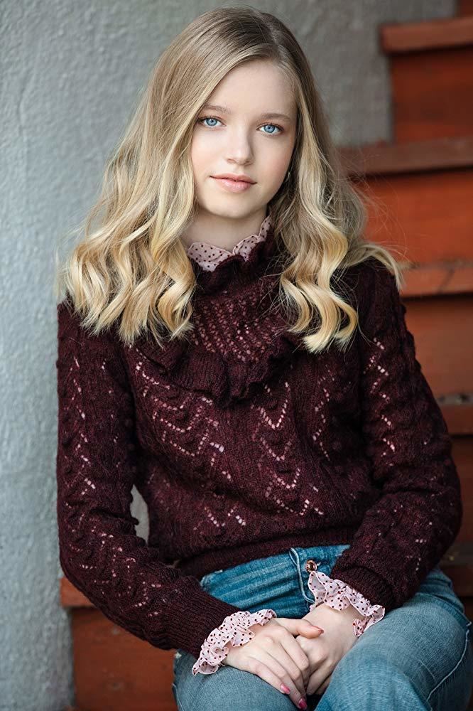 Shay Rudolph with a tight-lipped smile while sitting on the stairs and wearing a maroon crochet long sleeve blouse and denim pants