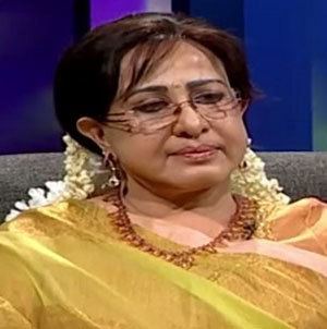 Sheela sitting on the chair during an interview while wearing orange dresses, yellow dupatta, gajra, eyeglasses, and some jewelry