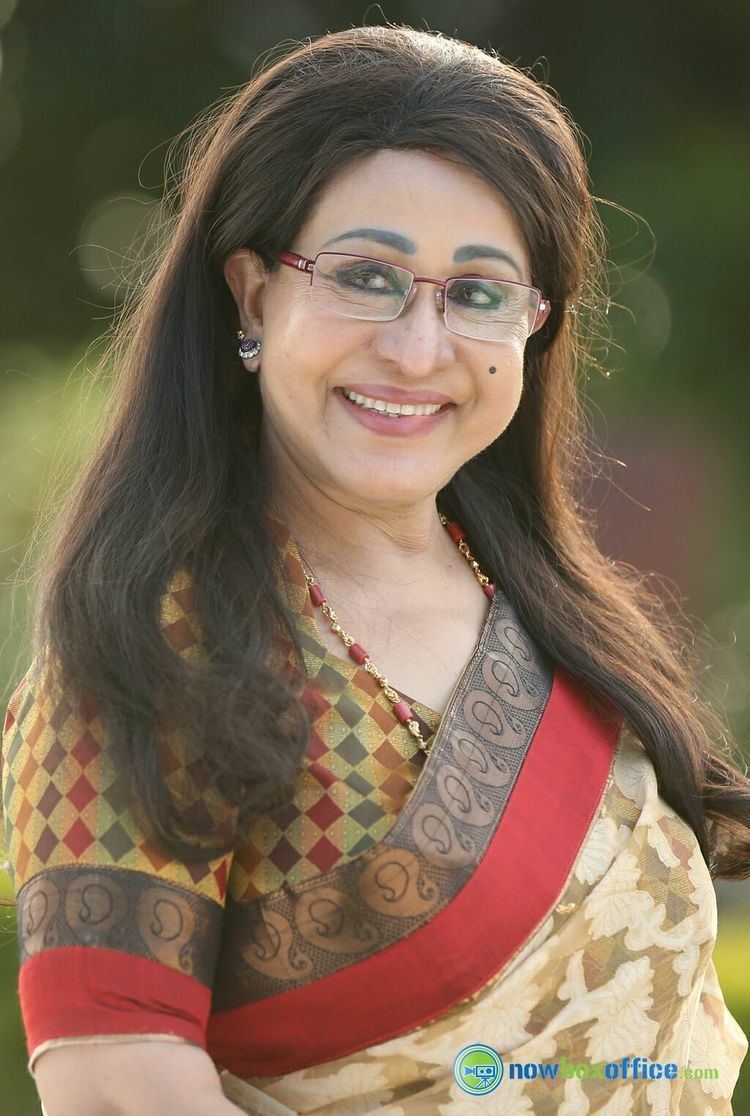 Sheela smiling while wearing a yellow, orange, and brown dress, eyeglasses, earrings, and necklace