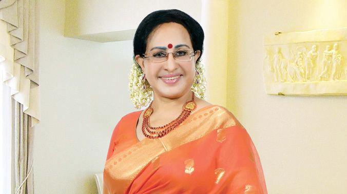 Sheela smiling while wearing an orange dress, gold and orange dupatta, necklace, eyeglasses, and a gajra on her hair