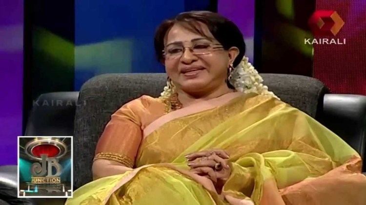 Sheela sitting on the chair during an interview while wearing an orange dress, a yellow dupatta, eyeglasses, and a gajra on her hair