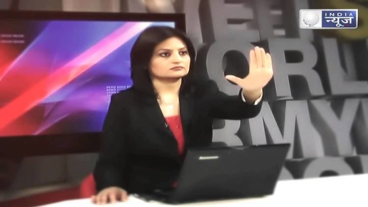 Sheetal Rajput reporting the news while extending her right arm and palm facing forward with a serious face, a laptop on the table and a tv monitor at the back, and she is wearing a red blouse under a black coat