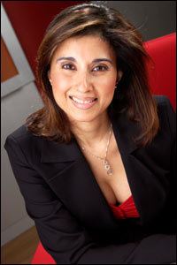 Shefali Oza smiling while sitting on a red chair and wearing a black coat, red inner blouse, and necklace