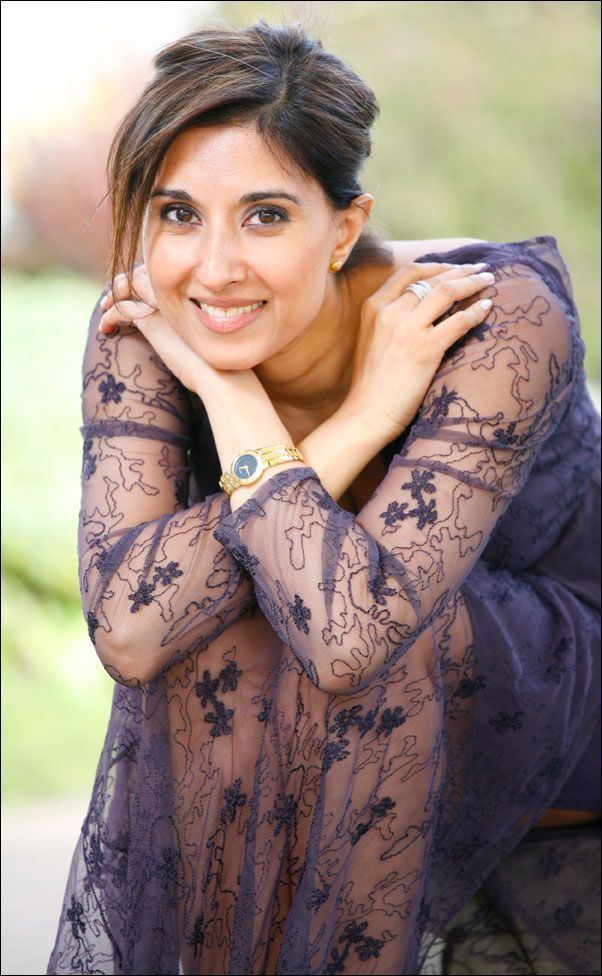 Shefali Oza smiling with her hands on her shoulder and wearing a blue lace long sleeve dress, wristwatch, ring, and earrings