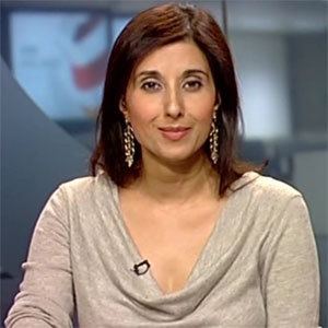 Shefali Oza with a tight-lipped smile while wearing a gray long sleeve blouse and earrings