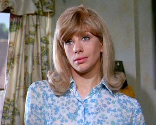 Sheila Fearn looking at something with a snobbish look with a cream and green curtain in the background. She has blonde shoulder-length curled hair and bangs, wearing a blue floral collared blouse.