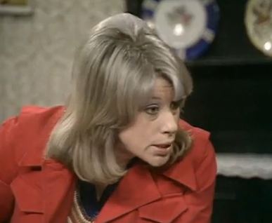Sheila Fearn talking to someone with a serious face and blonde shoulder-length hair and a fringe with a shelf and plates in the background, as Ann Fourmile in a 1980 Thames Television sitcom, "George and Mildred". She is wearing a brown and navy blue blouse under a red-orange blazer.